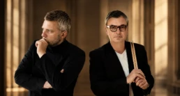 Conductor Kyril Karabits stands with percussion soloist Dimitris Desyllas in a refined editorial portrait for the Athens State Orchestra's performance of Timpani and Orchestra. Desyllas holds a pair of timpani mallets while both musicians are photographed in a warm contemporary concert hall interior.