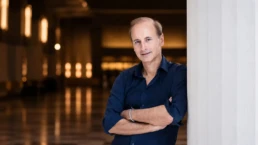 Conductor Miltos Logiadis in an editorial portrait for the Athens State Orchestra's performance of Mozart's Requiem, standing in an elegant concert hall interior with a warm, softly blurred background.
