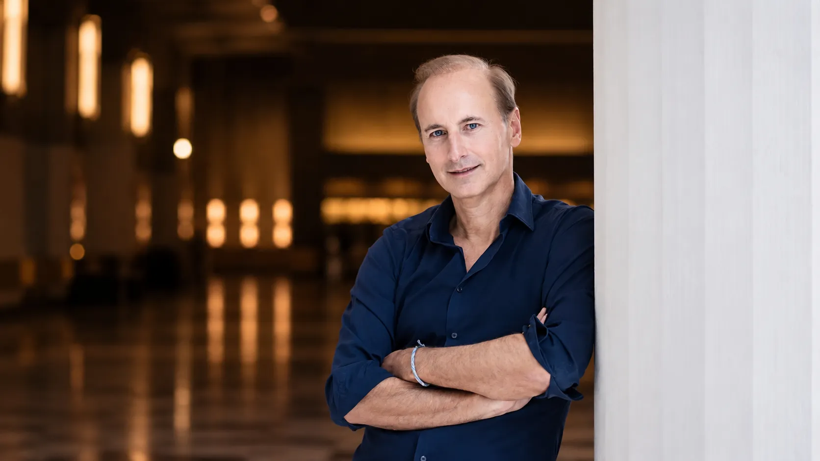 Conductor Miltos Logiadis in an editorial portrait for the Athens State Orchestra's performance of Mozart's Requiem, standing in an elegant concert hall interior with a warm, softly blurred background.