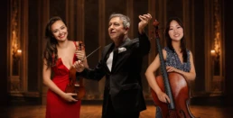 Conductor Loukas Karytinos stands at the center with baton raised, joined by a smiling cellist on the left and a violinist in a red dress on the right. The three musicians are posed together in a warmly lit concert hall with a dark theatrical background.