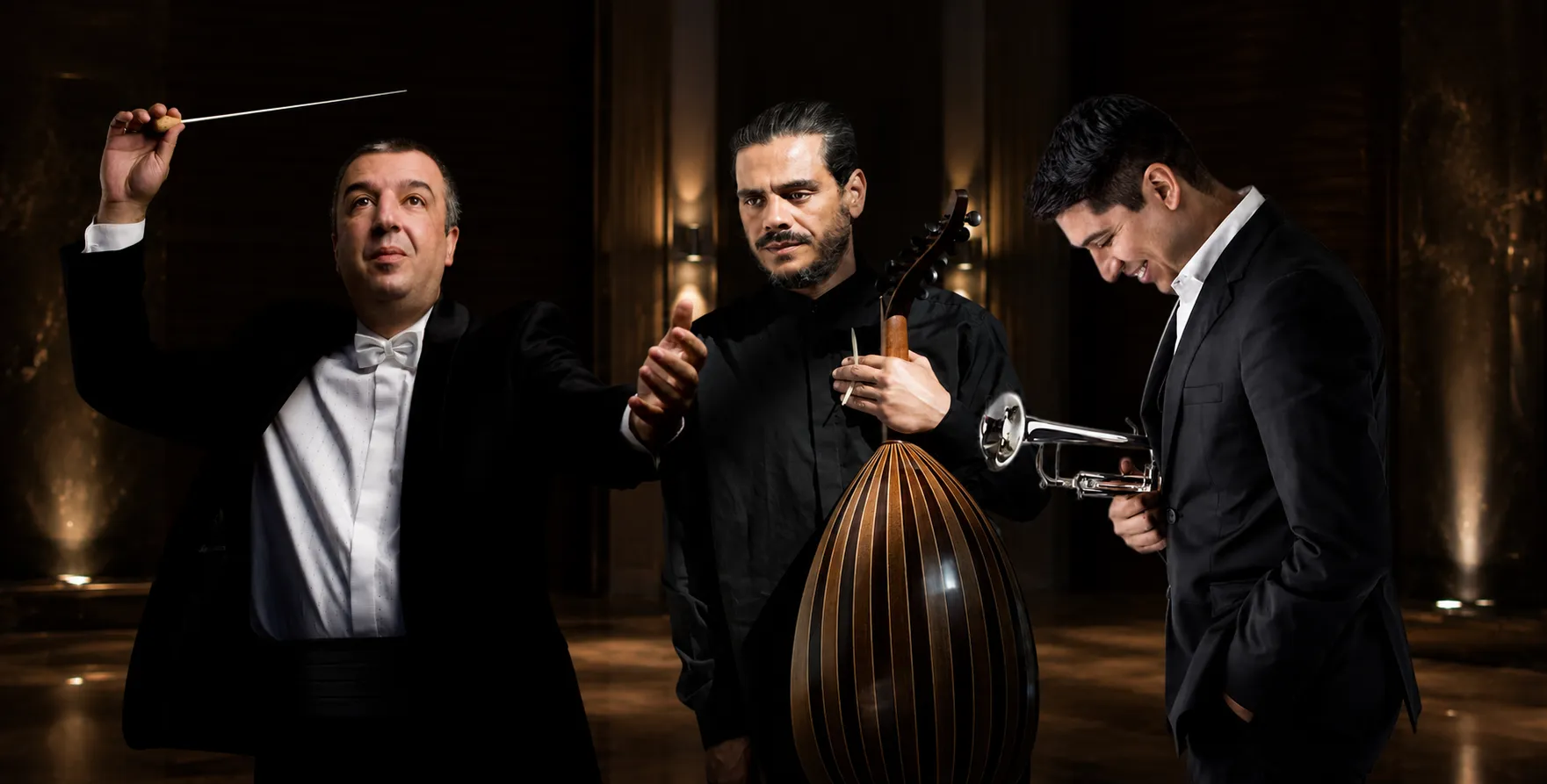 Three Athens State Orchestra musicians pose together inside an elegant concert hall. The conductor stands on the left with a raised baton, the oud soloist is at the centre holding his instrument, and the trumpeter stands on the right holding his trumpet. Warm theatrical lighting and the grand concert hall setting create the impression of a single professionally photographed ensemble.