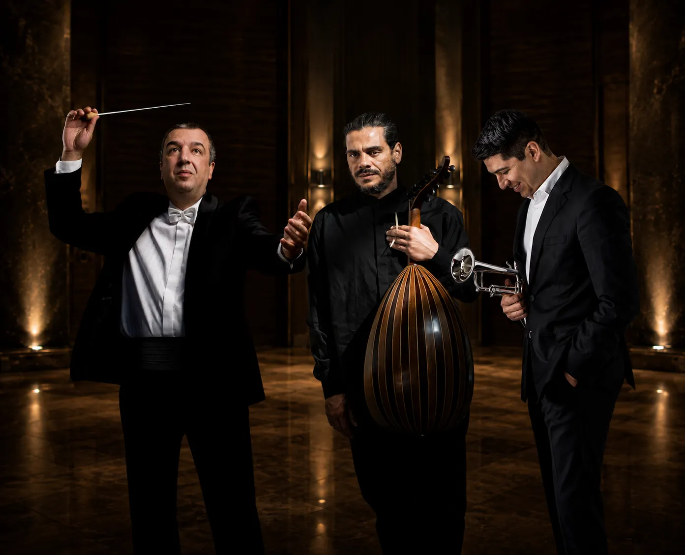 Three Athens State Orchestra musicians pose together inside an elegant concert hall. The conductor stands on the left with a raised baton, the oud soloist is at the centre holding his instrument, and the trumpeter stands on the right holding his trumpet. Warm theatrical lighting and the grand concert hall setting create the impression of a single professionally photographed ensemble.