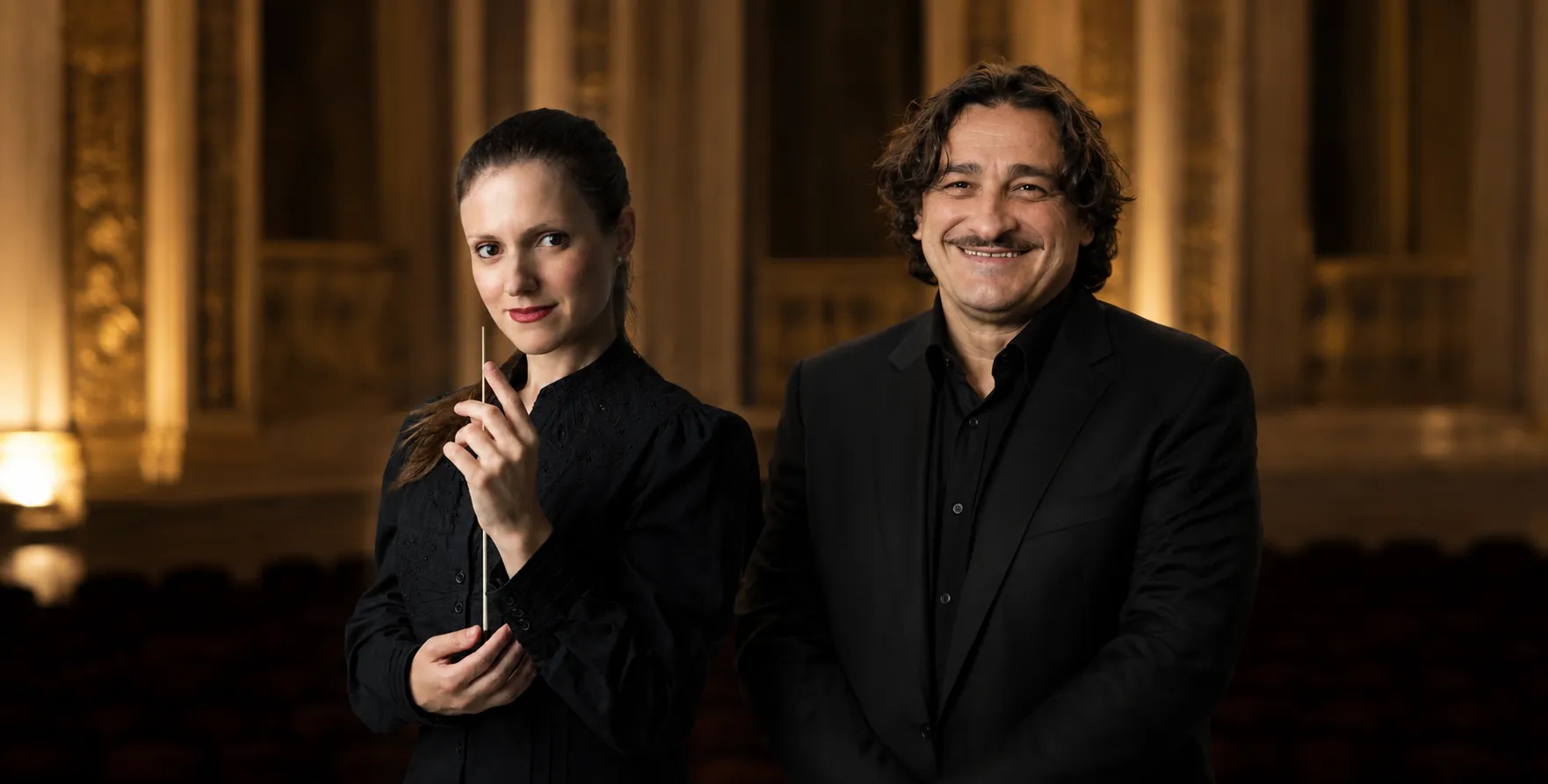 Premium editorial group portrait featuring conductor Myrsini Chatzikostanti and actor Vasilis Charalampopoulos, photographed in elegant formal attire with warm theatrical lighting and a refined concert-hall atmosphere.
