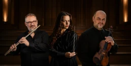 Conductor Maria Bazou stands between flutist Nikos Nikopoulos and violinist Apollon Grammatikopoulos in a warm editorial portrait for the Athens State Orchestra's performance of Music from Carmen, photographed in a luminous classical interior.